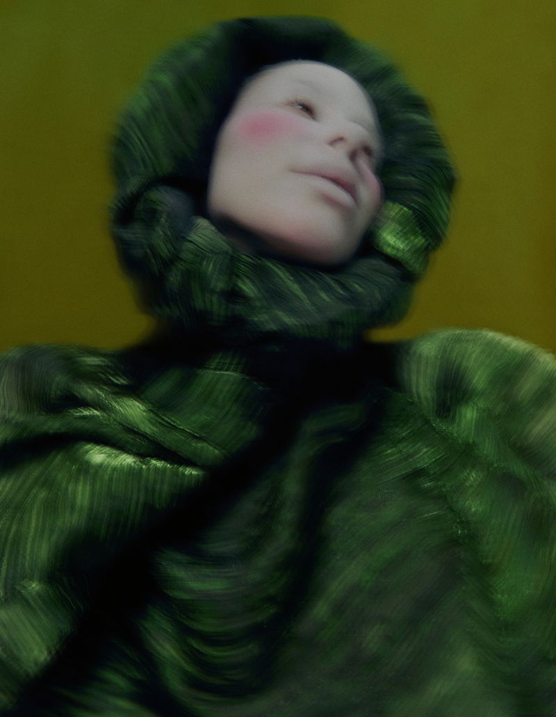 Close-up of a woman with a pale complexion wearing a hooded green garment, set against a muted yellow background in soft studio lighting.