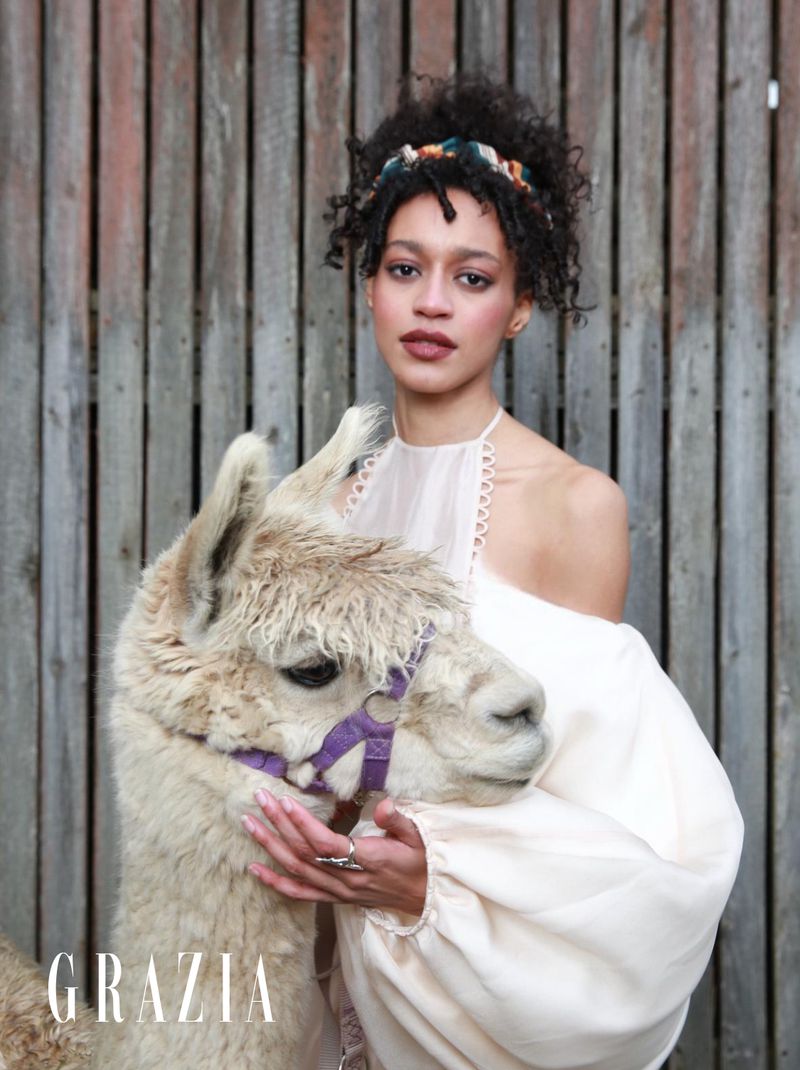 Portrait of a woman with curly hair wearing a light-colored dress, posing outdoors with an alpaca against a rustic wooden fence.