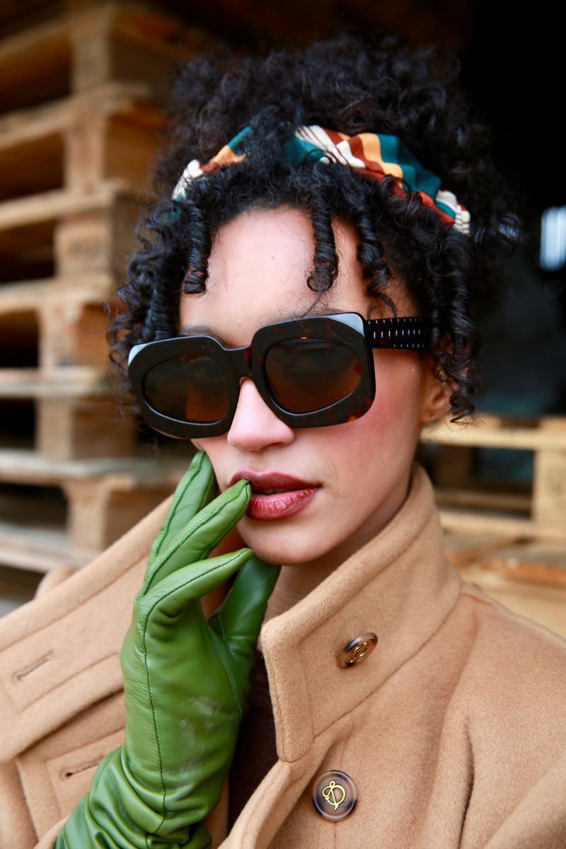 Fashionable adult female poses in a tan overcoat and large sunglasses with green gloves, set against a stacked wood pallet background.