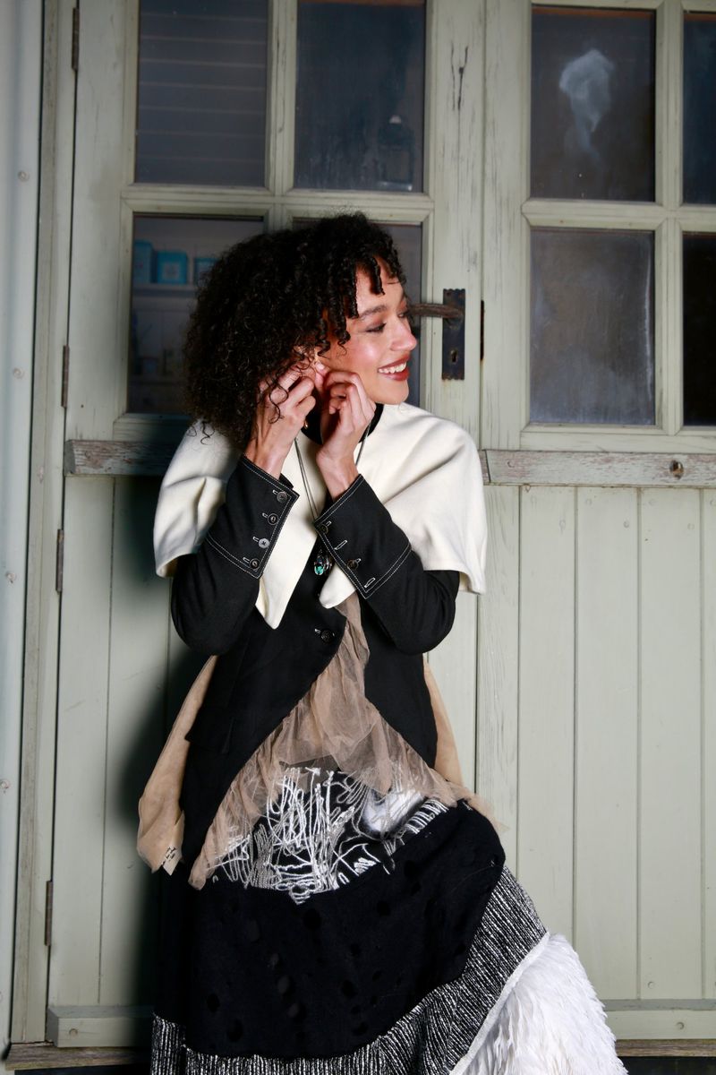 A woman in a black and white dress is adjusting her earrings in front of a weathered door with glass panels in natural light.