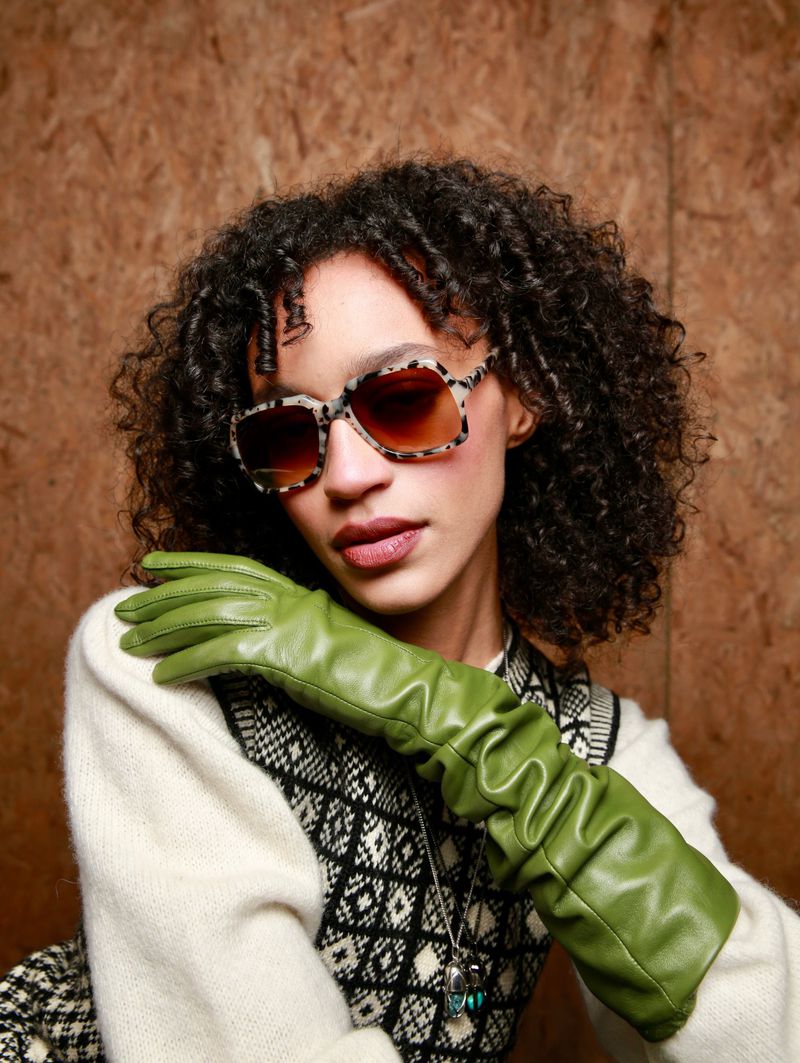 A brunette woman with curly hair and large sunglasses poses with long green gloves and a patterned sweater against a brown backdrop.