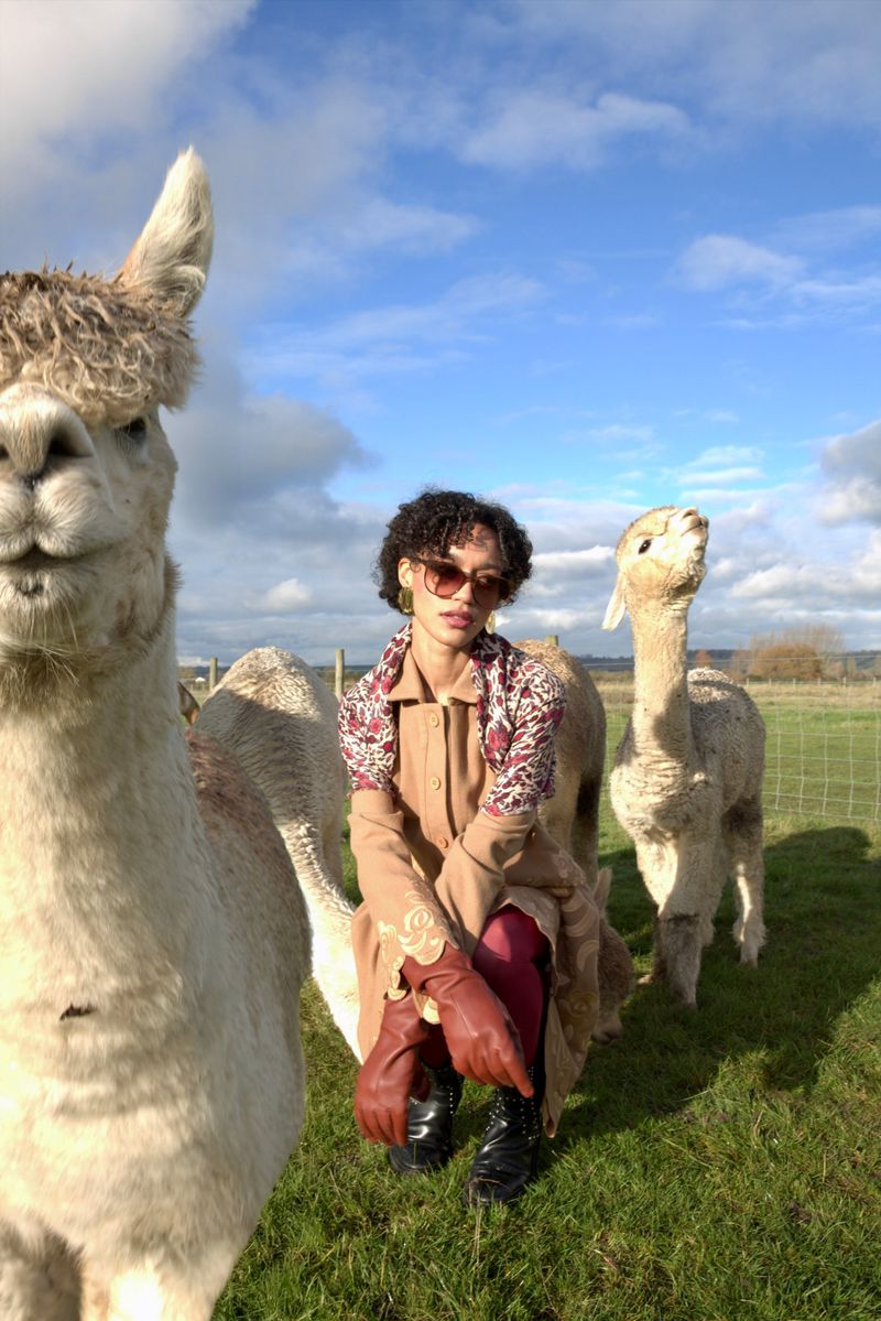 A young adult poses with three alpacas in an outdoor grassy field beneath a partly cloudy blue sky, showcasing a unique blend of fashion.
