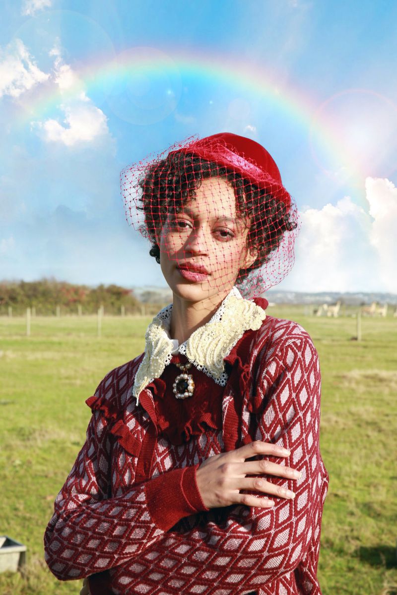 Portrait of a woman with short curly hair wearing a red hat and a red patterned sweater in a field under a rainbow sky.