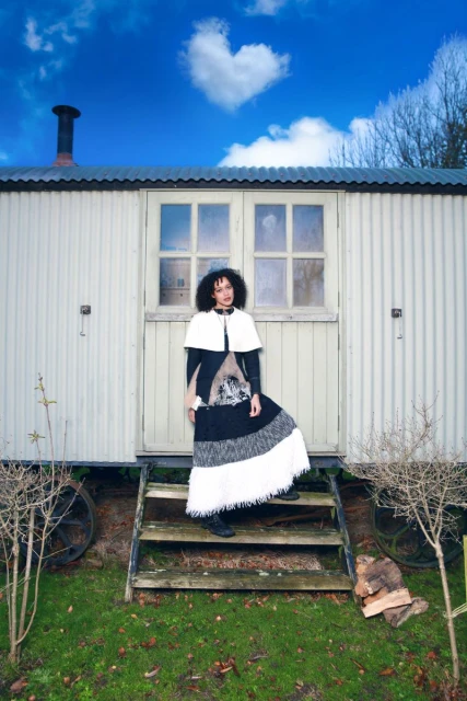 Adult woman stands on steps of white corrugated hut in a long patchwork dress and short white cape with blue sky and heart shaped cloud above.