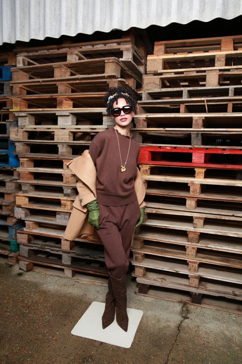 A stylish woman poses in a brown knit outfit, accessorized with sunglasses and a necklace, against a background of stacked pallets.