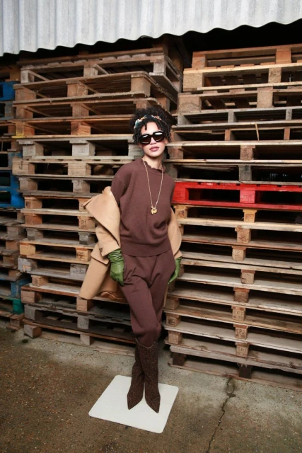 A stylish woman poses in a brown knit outfit, accessorized with sunglasses and a necklace, against a background of stacked pallets.