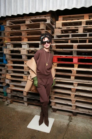 A stylish woman poses in a brown knit outfit, accessorized with sunglasses and a necklace, against a background of stacked pallets.