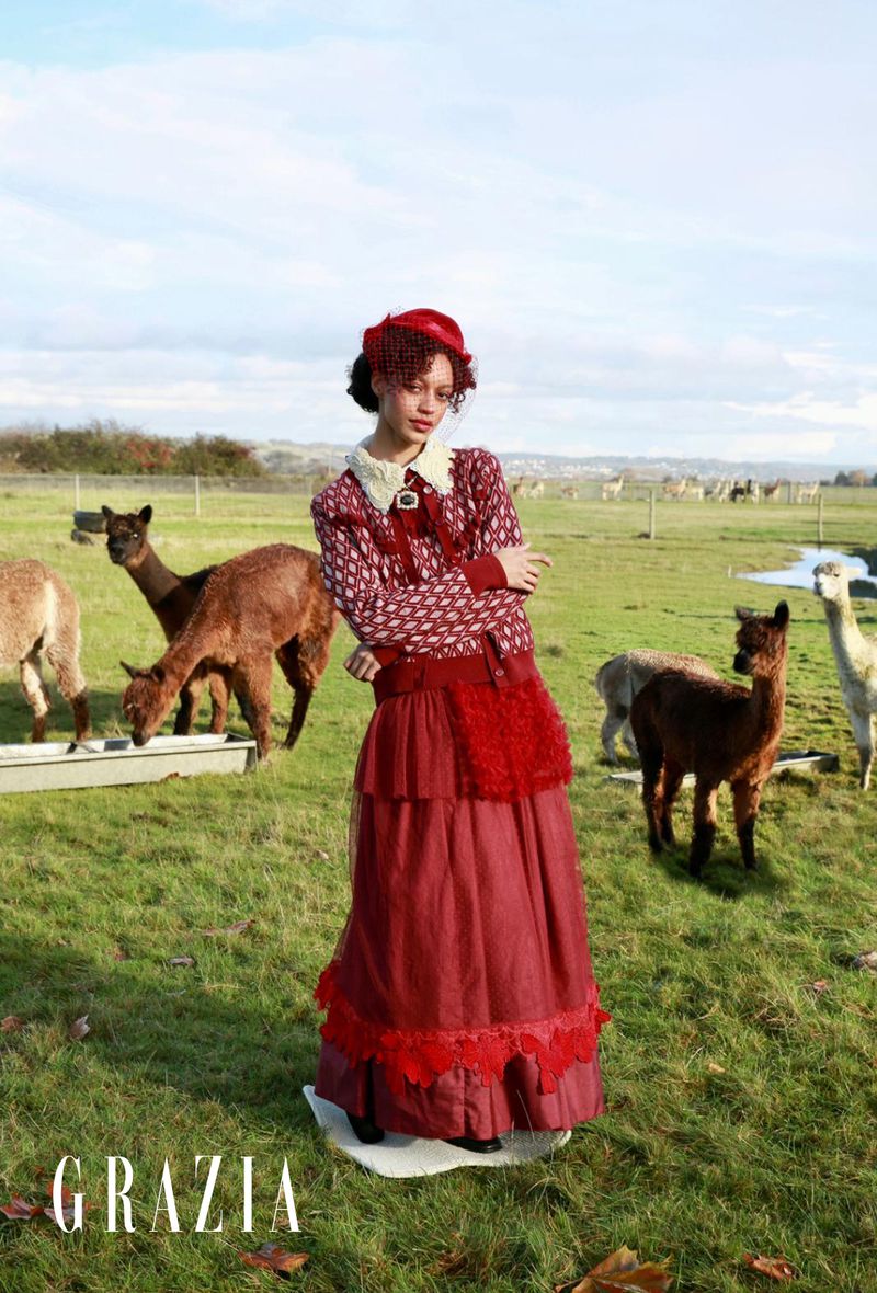 A young woman in a vintage-style dress stands in a grassy field surrounded by alpacas, under a bright sky with scattered clouds.