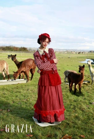 A young woman in a vintage-style dress stands in a grassy field surrounded by alpacas, under a bright sky with scattered clouds.
