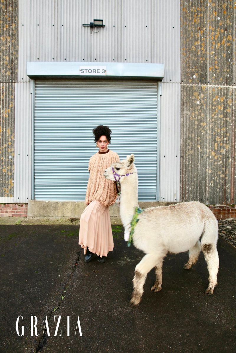 A fashionable adult woman with curly hair stands next to an alpaca in an outdoor setting; metal building and Grazia logo in view.