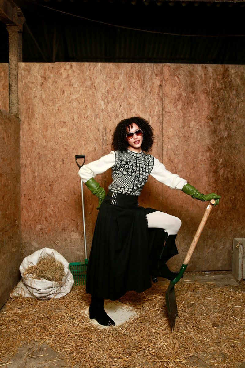 A person with curly hair and sunglasses poses in a barn with a shovel, wearing gloves, boots, and a long black skirt for a fashion shoot.