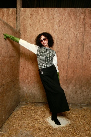 Model in long green gloves, patterned sweater vest, and black maxi skirt posing against plywood walls with straw flooring.