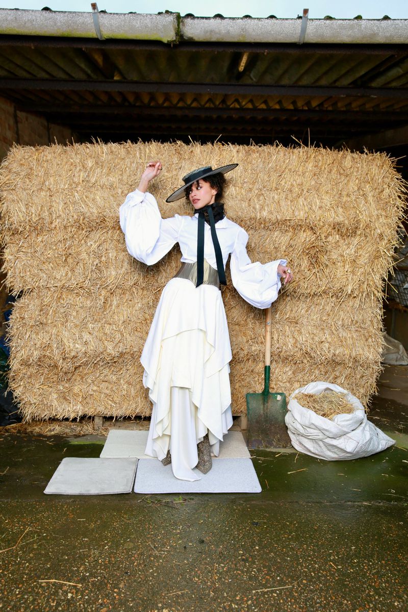 A woman poses in a white dress, top, and hat, standing against a backdrop of hay bales in a rural, outdoor setting with soft, diffused lighting.