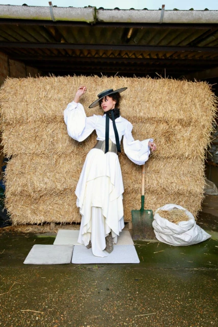 A woman poses in a white dress, top, and hat, standing against a backdrop of hay bales in a rural, outdoor setting with soft, diffused lighting.