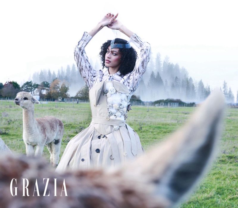 Fashion editorial photo of a woman in a beige gown standing in a field with an alpaca, set against a misty forest backdrop.