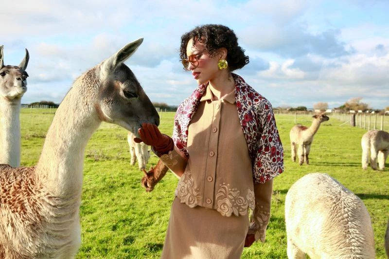 A woman in vintage fashion petting a llama in a grassy field under a blue sky with other llamas grazing in the background.