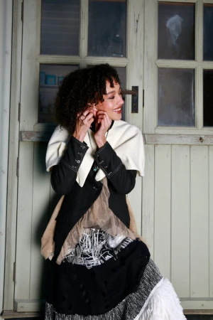 Smiling woman with curly hair wearing a layered black jacket and tiered skirt standing against a paneled wooden door.