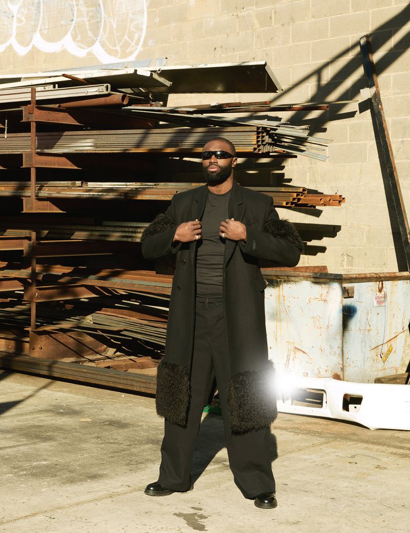 Adult male in a black coat with fur accents poses in an urban setting with stacked metal and a neutral concrete background on a sunny day.