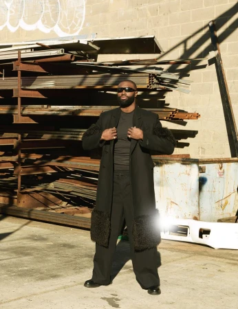 Adult male in a black coat with fur accents poses in an urban setting with stacked metal and a neutral concrete background on a sunny day.