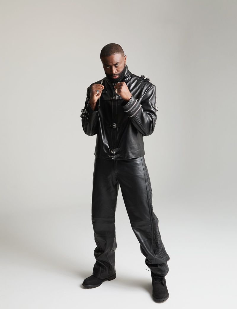 Portrait of a Black man posing in a black leather jacket and pants against a light gray backdrop, fists up in a boxing pose.