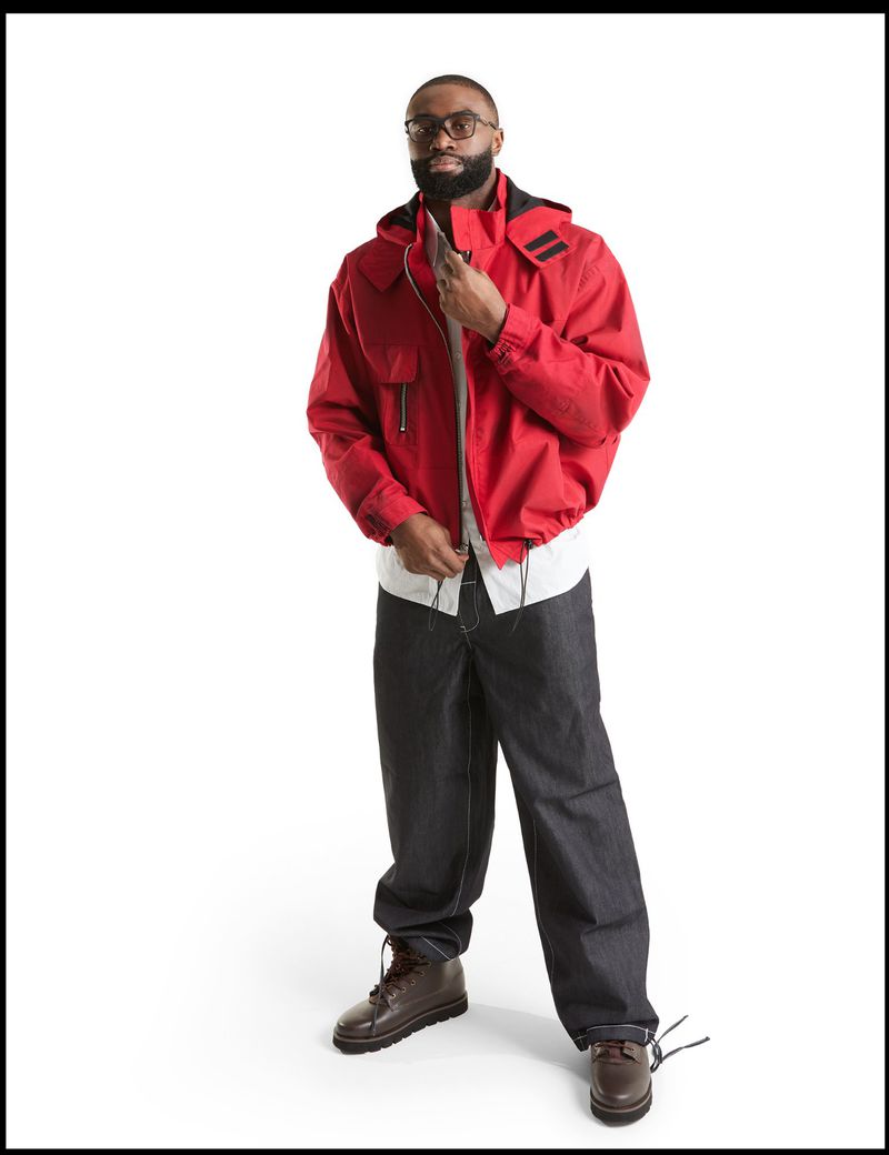 Full body studio shot of an adult male with a beard wearing a red hooded jacket, jeans, and boots against a white backdrop.