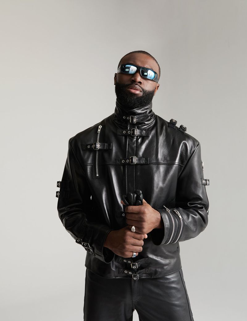 Jaylen Brown wearing a black buckled leather jacket and futuristic sunglasses against a light grey seamless backdrop.