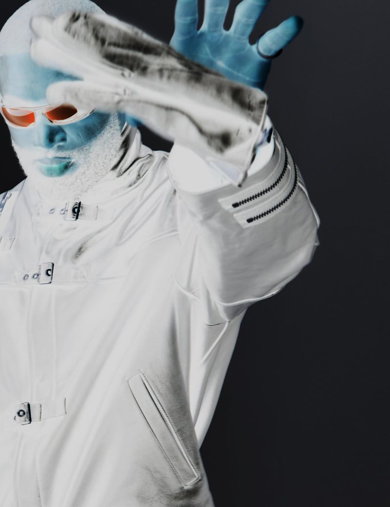 Stylized portrait of a man in a white coat and orange glasses raising his hand in a studio with contrasting colors.