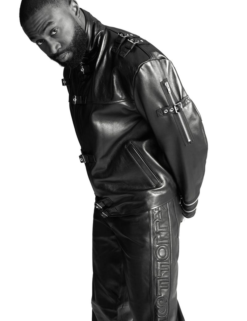 A stylish adult male in a sleek black leather jacket and matching pants, looking intently at the camera against a crisp white backdrop.