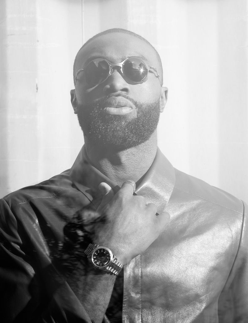 Black and white portrait of Jaylen Brown wearing round sunglasses, a leather jacket, and a watch while adjusting his collar.