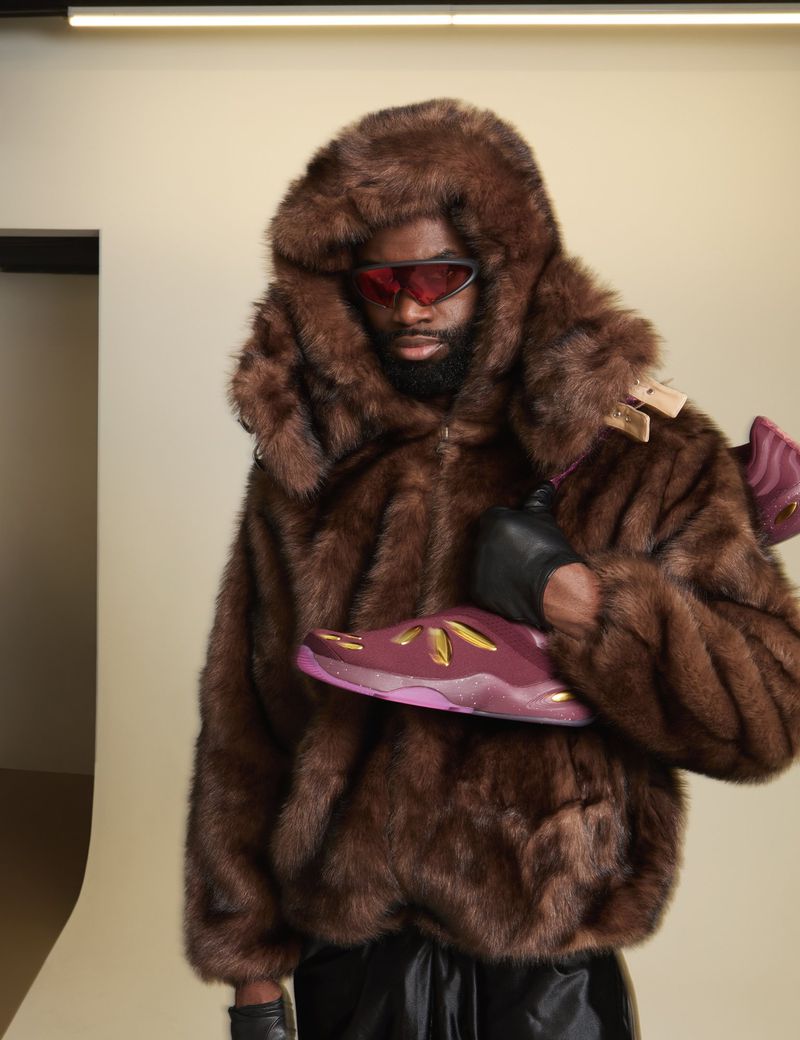Person wearing a fur coat with sunglasses, holding a sneaker. Indoor studio shot featuring a brown fur coat and futuristic sunglasses.
