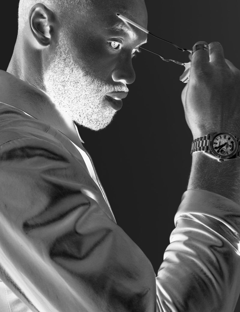 Monochrome portrait of a dark-skinned adult male indoors, wearing a collared shirt and a watch, using scissors, against a dark background.