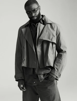 Black man with a beard wearing sunglasses, a collared shirt, a cropped jacket, and trousers against a grey backdrop