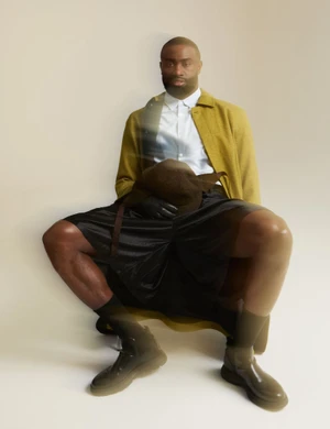 Jaylen Brown seated wearing a yellow jacket, white shirt, and black shorts against an off-white studio backdrop.