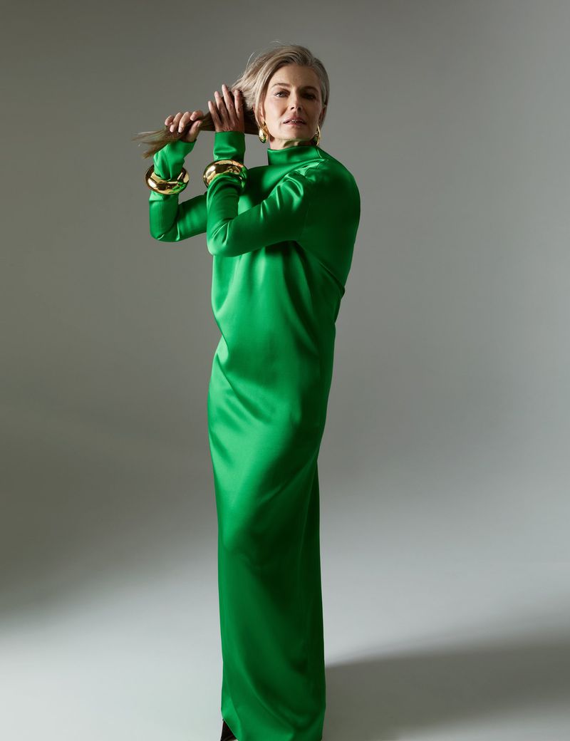 A woman in a green dress stands in a studio, holding her hair up with her hands and looking at the camera with a neutral expression.