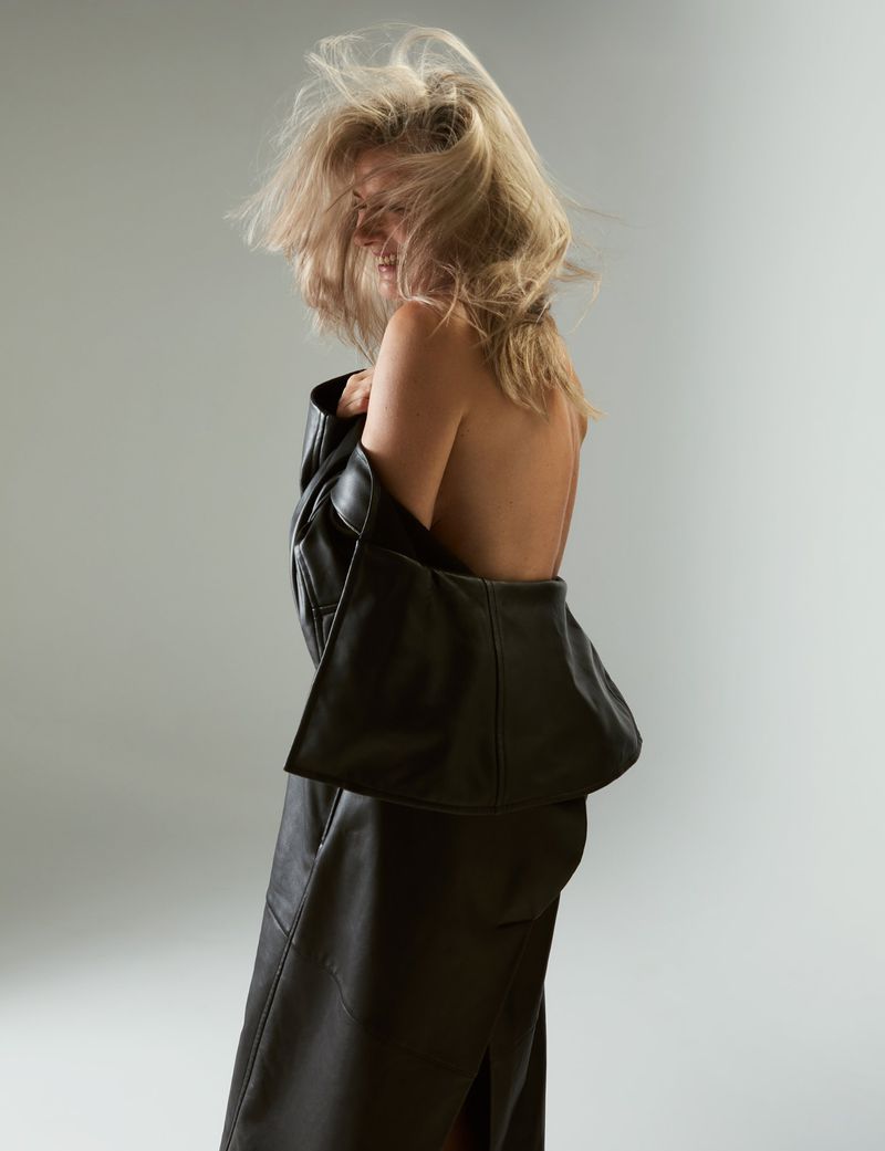 Blonde woman poses in a dark leather coat in a studio environment. Back shot of a fashion model with wind-blown blonde hair.