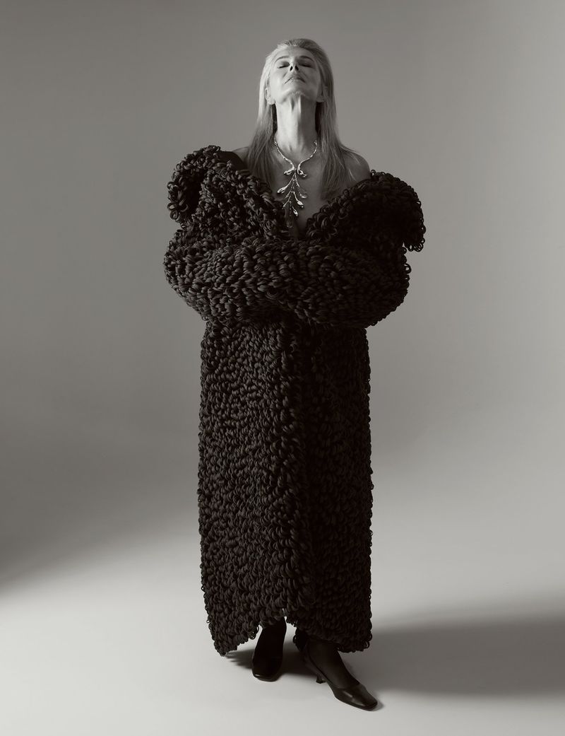 Elegant mature woman poses in a studio wearing a textured black coat and statement necklace with her head tilted back and eyes closed.