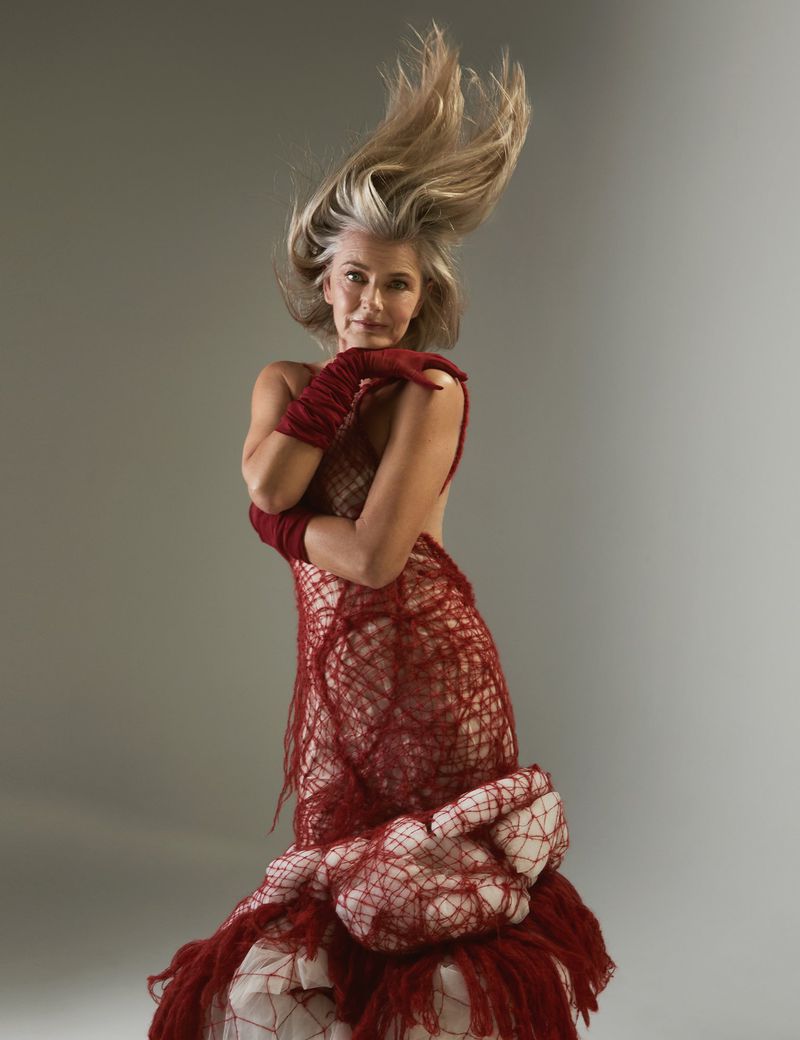 Older adult female model with grey hair and a red textured couture dress poses in a studio with arms crossed and hair blowing upward.