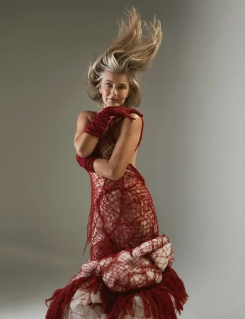Older adult female model with grey hair and a red textured couture dress poses in a studio with arms crossed and hair blowing upward.