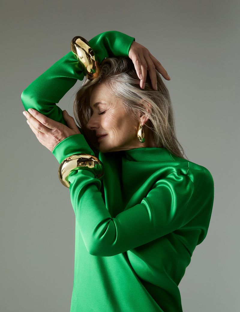 Elegant older woman in green dress poses with gold jewelry. Fashion portrait in studio with soft lighting and neutral background.