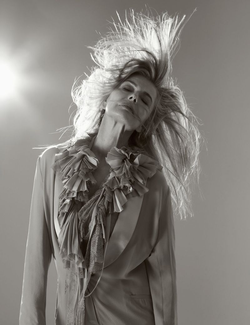 Monochrome portrait of a woman with long blonde hair in motion wearing a ruffled jacket against a pale gray background with a light.