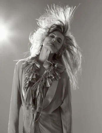 Monochrome portrait of a woman with long blonde hair in motion wearing a ruffled jacket against a pale gray background with a light.