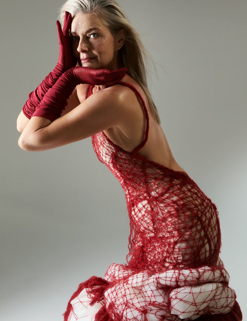 An older woman poses in a studio wearing a red knit dress and gloves, looking at the camera with a serene expression.