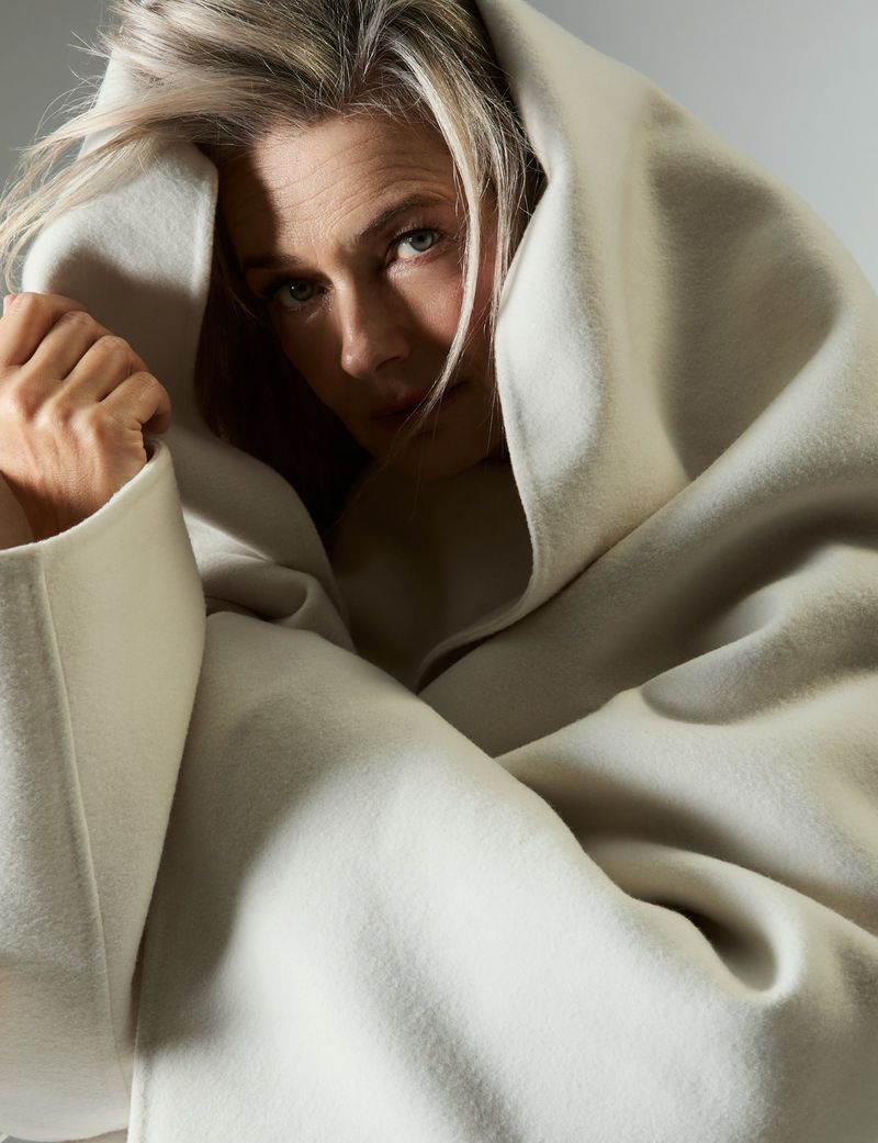 Close-up studio portrait of a woman with blonde hair wrapped in a soft, light-beige wool coat with a hood, gazing directly at the camera.