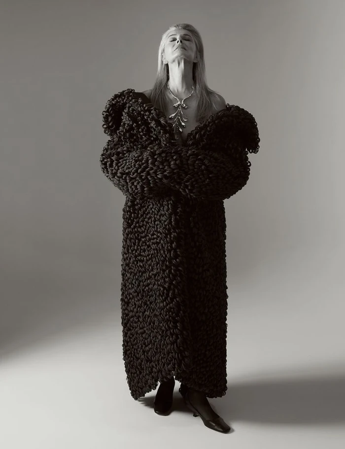 Model Paulina Porizkova wearing a voluminous black textured knit maxi coat and statement necklace against a grey studio backdrop.