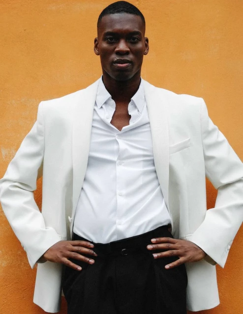 A fashionable Black man in a white jacket and black pants poses confidently against an orange wall, conveying style and sophistication.