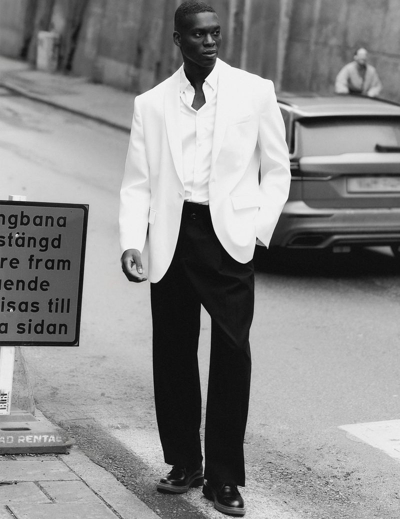 Black and white portrait of a Black male model wearing a white blazer and black pants, standing on a city street next to a road sign.