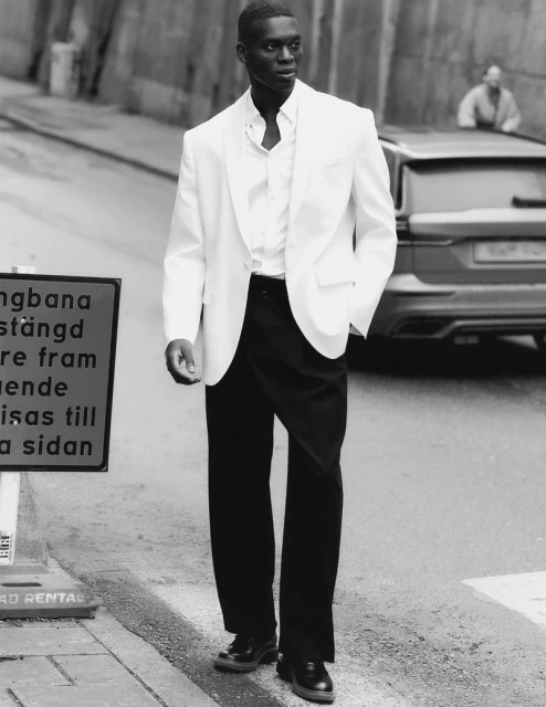 Black and white portrait of a Black male model wearing a white blazer and black pants, standing on a city street next to a road sign.