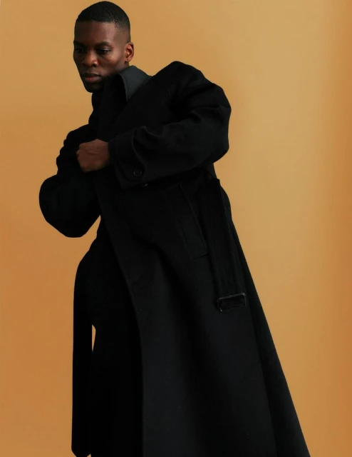 Portrait of a Black male model in a long, belted black coat, posing indoors against a neutral background, looking to the side.