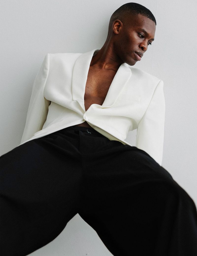 A Black male model poses in a white blazer and black wide-leg pants against a neutral background, captured in a medium shot with studio lighting.