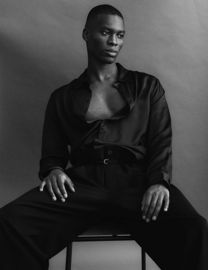 Black and white portrait of a Black male model with an unbuttoned silk shirt sitting on a stool in front of a gray background.